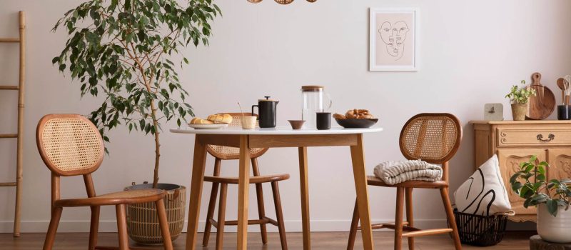 stylish-dining-room-with-round-table-rattan-chair-wooden-commode-poster-kitchen-accessories-beige-wall-with-mock-up-poster-home-decor-template